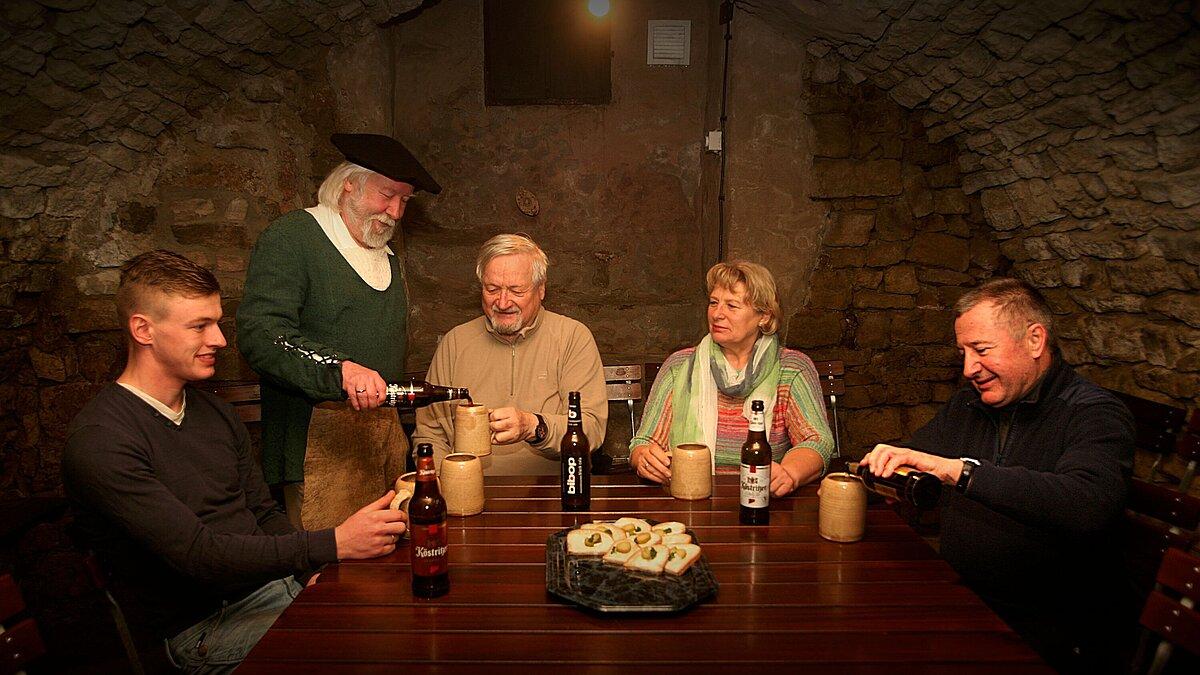Die Gäste sitzen in einem historischen Höhler. Ein Herr im historischem Kostüm schenkt den Gästen Bier ein. 